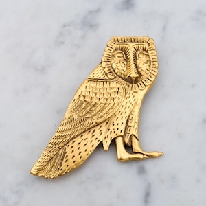 May include: A gold-toned metal brooch shaped like an owl, featuring intricate engraved feather patterns across its wings and body, a detailed facial disc, and a single visible leg extending from the bottom of the bird's body in a profile view.