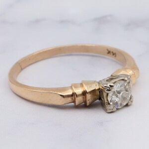 May include: A gold engagement ring featuring a single round diamond set in a prong setting. The band is a simple, thin band with a slightly textured finish. The ring is marked "14K" on the inside of the band.