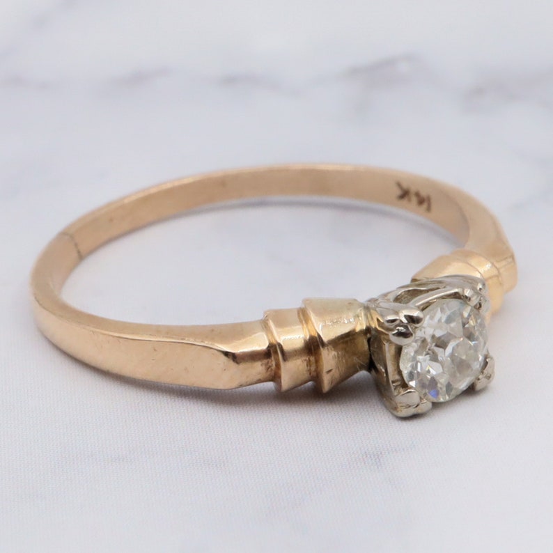May include: A gold engagement ring featuring a single round diamond set in a prong setting. The band is a simple, thin band with a slightly textured finish. The ring is marked "14K" on the inside of the band.