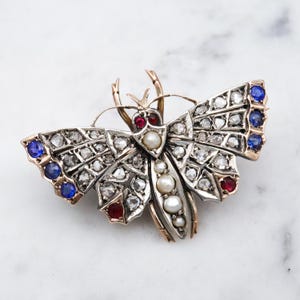 May include: A detailed brooch in the shape of a moth, featuring a silver-tone body with gold accents. The wings are adorned with clear and blue gemstones, along with red gems and pearls. The brooch is set against a white marble background.