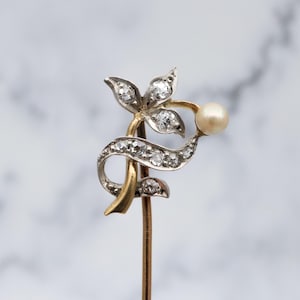 Könnte beinhalten: Vintage Hutnadel in Gold- und Silberfarben, mit floralem Design, besetzt mit klaren Edelsteinen und einer einzelnen Perle. Die Nadel hat einen schlanken, goldfarbenen Stiel und eine dekorative Oberseite mit Blumen- und Bandmotiv.