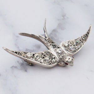 Puede incluir: Un broche de plata en forma de golondrina con detalles de pedrería transparente. Las alas de la golondrina están extendidas y el pájaro está mirando hacia la derecha. La golondrina tiene un strass rojo para el ojo.