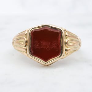 May include: A gold signet ring with a red carnelian stone set in a shield shape. The stone has an engraved design.