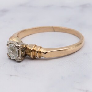 May include: A gold band engagement ring with a single round diamond set in a prong setting. The band is slightly thicker on one side, creating a subtle design detail.