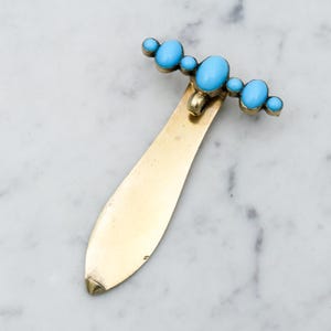 Puede incluir: Un abrecartas dorado vintage con una parte superior decorativa con cinco piedras ovaladas de turquesa azul claro. El abrecartas tiene una hoja larga y delgada y un pequeño lazo en la parte superior.