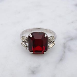 May include: A silver ring featuring a large, rectangular, deep red gemstone as the centerpiece. Smaller, clear rectangular stones flank the main stone. The ring is set against a white marble surface.