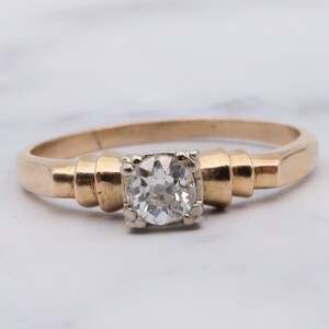 May include: A gold engagement ring featuring a single round diamond set in a white gold setting. The band is a simple, flat design with a slightly rounded edge.