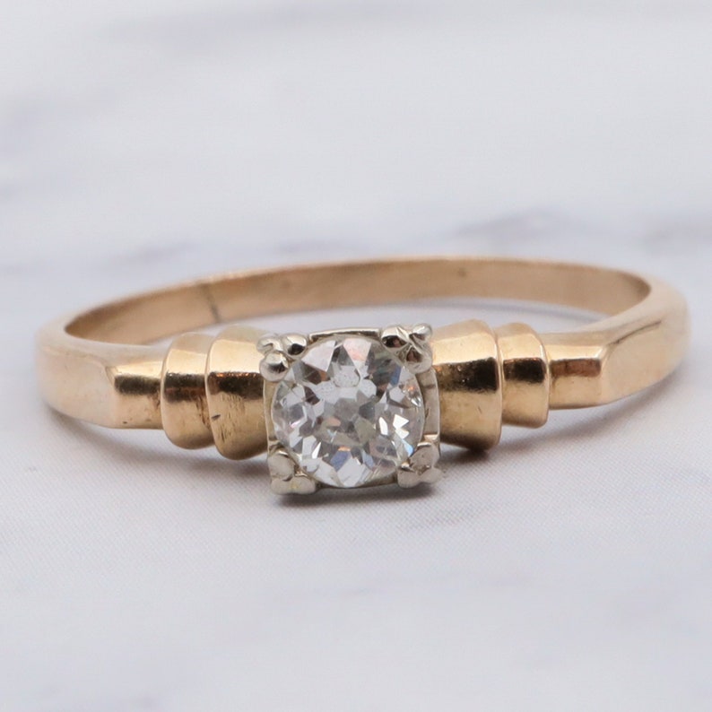 May include: A gold engagement ring featuring a single round diamond set in a white gold setting. The band is a simple, flat design with a slightly rounded edge.