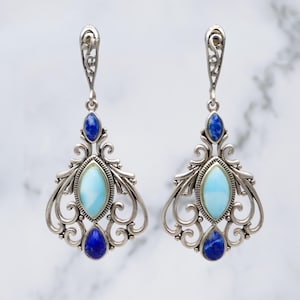 May include: A pair of silver-toned drop earrings. Each earring features a light blue oval stone, surrounded by a decorative silver frame, and accented with dark blue stones. The earrings have an ornate, filigree design.