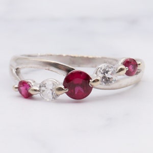 Könnte beinhalten: Ein silberner Ring mit einem gebogenen Band, besetzt mit drei roten Edelsteinen und zwei klaren Edelsteinen.