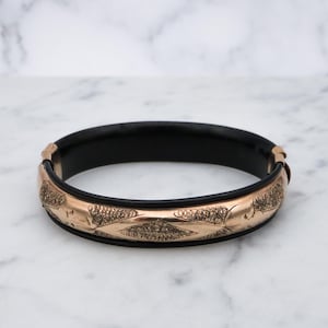Puede incluir: Una pulsera de brazalete con bisagras con interior negro y exterior dorado. El exterior dorado presenta un patrón en relieve de diseños estilizados de flores u hojas. La pulsera se exhibe sobre una superficie de mármol blanco.
