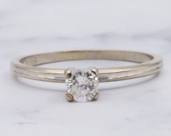 May include: A simple silver band ring with a single round diamond set in the center. The band has a textured design.