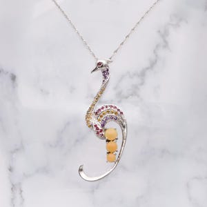 May include: A silver-toned necklace featuring an ostrich pendant. The pendant is adorned with colorful gemstones, including red, yellow, and purple stones, and three round, peach-colored stones. The necklace has a delicate chain.