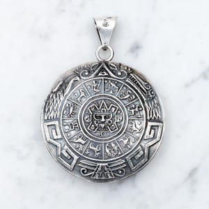 Puede incluir: Colgante plateado con un diseño circular detallado que presenta símbolos del zodíaco y una cara central. El colgante tiene un lazo para una cadena y probablemente está hecho de metal. El diseño es intrincado y parece hecho a mano.