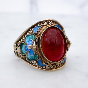 May include: A gold-toned ring featuring a large, oval-shaped, deep red cabochon stone at the center. The band is crafted with intricate filigree metalwork and decorated with vibrant blue and green enamel floral patterns surrounding the stone.