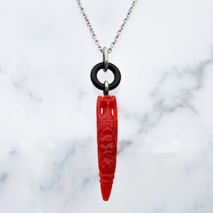May include: A silver chain necklace featuring a red, carved pendant in the shape of a tiki figure. The pendant is suspended from a black ring. The background is a white marble effect. The pendant has detailed carvings.