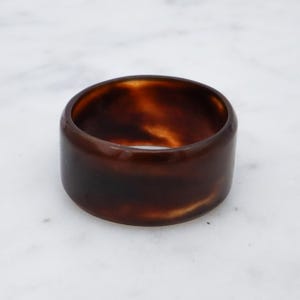 May include: A wide, brown, marbled bangle bracelet. The bracelet has a smooth, polished surface and a rounded shape. The color is a mix of dark and light browns, creating a tortoiseshell effect. The bangle is approximately 1 inch wide.