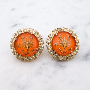 May include: A pair of vintage gold-toned earrings. Each earring features a round, orange center with a gold scorpion design and a ring of clear rhinestones. The earrings are set against a white marble background.