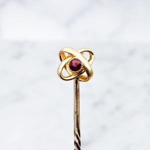 Puede incluir: Un prendedor de corbata de oro con una piedra preciosa roja en el centro. El prendedor tiene forma de nudo.