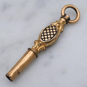 May include: A gold-toned pocket watch key with a black and gold checkered design on the top. The key has a loop at the top for attaching to a chain.