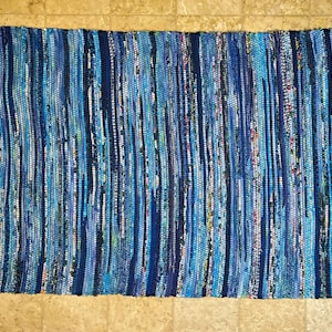 May include: A rectangular woven rag rug featuring a vertical striped pattern in various shades of blue, including navy, royal, and light blue. The rug has a textured, multi-tonal appearance with visible fabric strips and short fringe on both ends.