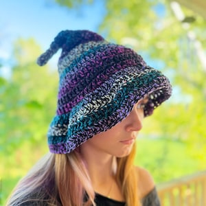 May include: A crocheted bucket hat featuring a pointed top and a wide, downward-sloping brim. The hat is crafted from variegated yarn in a dark colour palette, showcasing blended stripes of deep purple, black, teal, and silver-grey throughout the design.