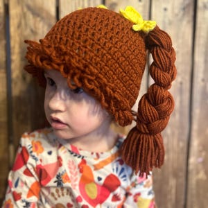 May include: A brown crocheted hat with a braided ponytail and yellow bows. The hat is worn by a child wearing a colorful shirt with a rooster pattern.