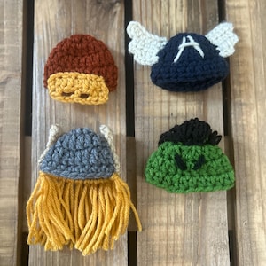 May include: Four crocheted hats, each representing a different superhero. The hats are brown, blue, gray, and green. The blue hat has white wings and the letter "A" on it. The gray hat has a yellow beard. The green hat has a green face with black eyes.