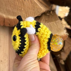 May include: A handmade, crocheted bee toy in yellow and black stripes, with a white patch and black eyes. The bee has a clear, glitter-filled orb at the end. The toy is held in a hand against a wood background.