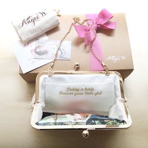 Puede incluir: Un bolso de mano de sat&eacute;n blanco con cierre dorado y cadena dorada. El bolso est&aacute; abierto y el forro interior es blanco con bordado dorado que dice "Today a bride, forever your little girl". El bolso est&aacute; empaquetado en una caja marr&oacute;n con una cinta rosa.