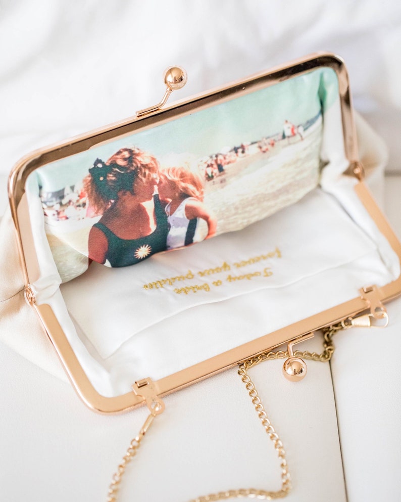 Puede incluir: Un bolso de mano dorado con una cadena. El bolso est&aacute; forrado con tela blanca y tiene una foto de dos personas en la playa impresa en la parte delantera. El interior del bolso tiene el texto "2. Coconut or Bride" y "Forever yours darling" bordado en hilo dorado.