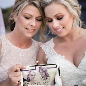 A bride with her mother on the wedding day with both holding and looking down smiling at a personalized photo bag showing a photo of them together.  Bride in her bridal gown and the mother of the bride is wearing a champagne beaded dress.