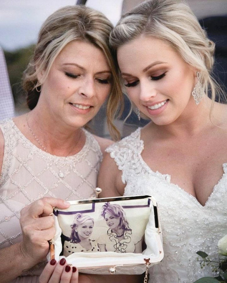 A bride with her mother on the wedding day with both holding and looking down smiling at a personalized photo bag showing a photo of them together.  Bride in her bridal gown and the mother of the bride is wearing a champagne beaded dress.