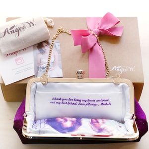 Photo of the inside of a purple clutch bag showing a personalized photo and a custom 2-line embroidered message in purple.  The bag is propped against a branded ANGEE W. brown kraft paper box with pink ribbon, a dust bag and thank you card.