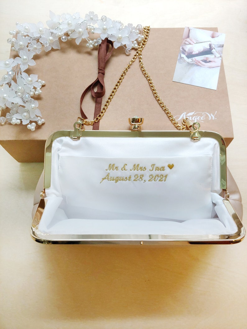Puede incluir: Un bolso de mano dorado con forro de sat&eacute;n blanco y una cadena dorada. El bolso tiene una inscripci&oacute;n personalizada que dice "Mr. & Mrs. Ina August 28, 2021" con un peque&ntilde;o coraz&oacute;n.