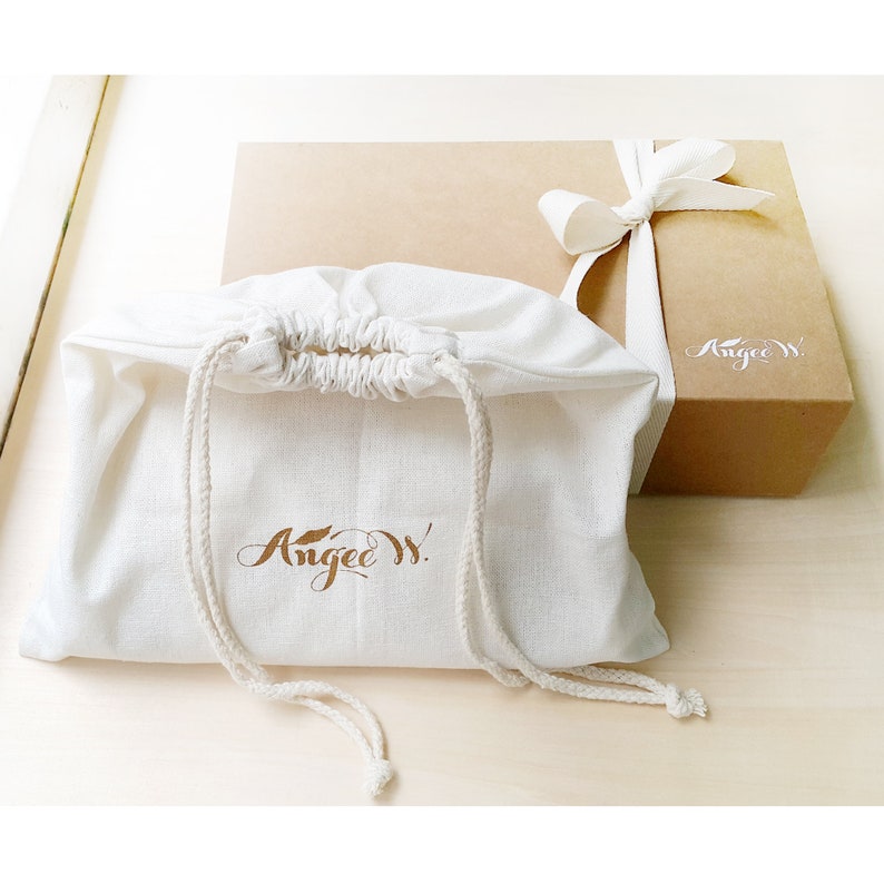 Image showing a clutch bag inside a dust bag leaning against an ANGEE W. branded brown kraft paper gift boxes with an ivory ribbon