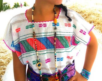 May include: A white embroidered tunic with colorful geometric patterns, a straw hat, and beaded jewelry. The tunic has a pink trim and is paired with a floral belt and a beaded bracelet. The overall style is bohemian.