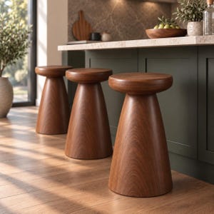 May include: Three solid wood stools with a smooth, rounded seat and a tapered, conical base. The stools feature a rich, warm brown wood grain finish and are positioned in a row on a wooden floor, showcasing a consistent, sturdy design.