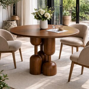 Puede incluir: Una mesa de comedor redonda de madera con una base de pedestal única, rodeada de cuatro sillas tapizadas beige. Un jarrón blanco con flores está sobre la mesa, junto con una taza y material de lectura. La mesa tiene unos 122 cm de diámetro.