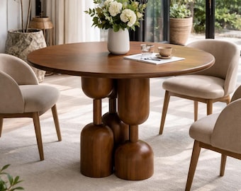 May include: A round wooden dining table with a smooth surface and a base composed of several stacked, rounded, bulbous wooden pillars. The table is surrounded by four beige upholstered chairs with wooden legs, arranged on a light-coloured rug.