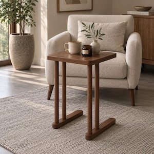 May include: A square wooden side table with four cylindrical legs and two horizontal base supports. The table surface is dark brown and holds a beige ceramic mug and a small dark brown glass jar with a label.