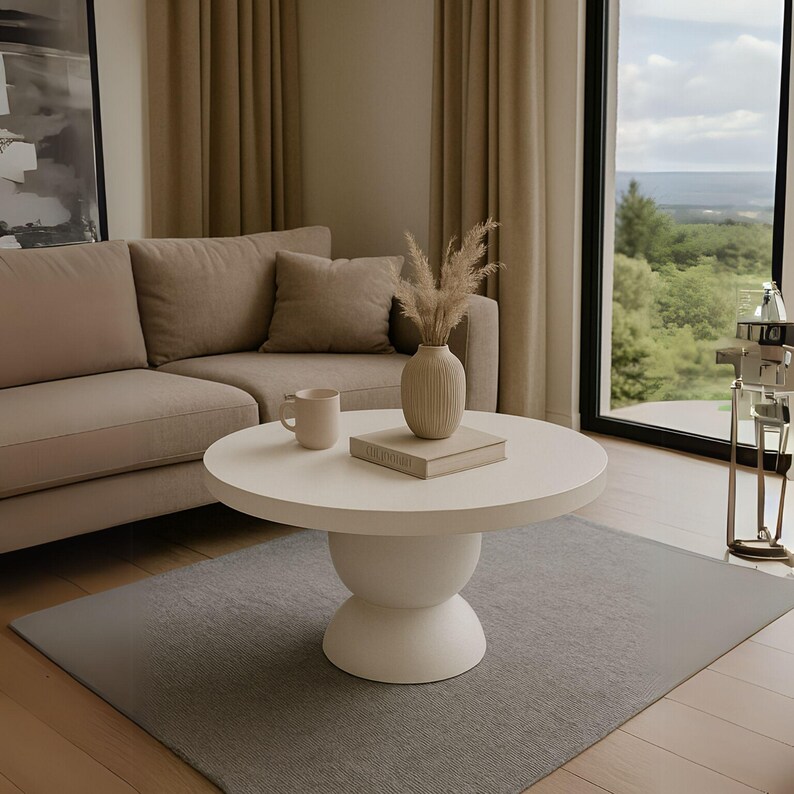 May include: A white round coffee table with a pedestal base sits on a gray rug in a living room. A beige sofa with pillows is in the background. A vase with dried pampas grass and a book are on the table.