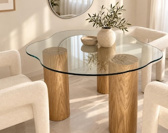 May include: A dining table featuring a clear, organic-shaped glass top supported by three cylindrical light wood legs with a visible grain. A textured beige ceramic vase containing green leafy branches sits on the glass surface.