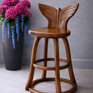 May include: A wooden bar stool with a butterfly wing-shaped backrest. The stool is made of light brown wood and features a round seat and a circular footrest. The legs are curved and the stool is placed in front of a gray wall.