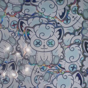 May include: A collection of stickers featuring a light blue plushie-like creature with button eyes and a stitched design. The stickers have a glittery, iridescent border and a cloud-like shape.