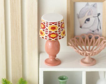 May include: Miniature table lamp with a peach-colored base and a patterned lampshade. The lampshade features a geometric design in red, yellow, and white. The lamp is part of a dollhouse decor set, alongside a framed picture and a decorative bowl.