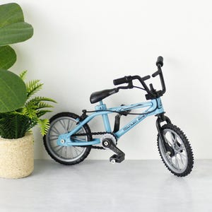 Könnte beinhalten: Ein hellblaues Miniatur-Mountainbike mit schwarzen Lenkern, Sattel und Reifen. Das Fahrrad hat eine silberne Kette und Pedale. Die Räder sind silberfarben mit schwarzen Reifen. Das Fahrrad steht auf einer weißen Oberfläche.
