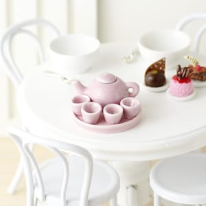 May include: A pink porcelain tea set with a teapot and five cups on a white saucer. The tea set is on a white table with two white chairs.
