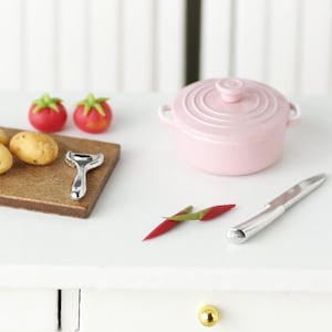 Könnte beinhalten: Ein rosa Keramiktopf mit weißen Griffen, ein Holzbrett mit einem silbernen Kartoffelschäler, zwei rote Tomaten, zwei rote Chilischoten und ein silbernes Messer auf einer weißen Arbeitsplatte.