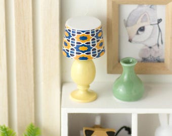 May include: A miniature yellow table lamp with a patterned lampshade featuring blue, orange, and white geometric designs. The lamp sits on a white shelf with a green vase and a framed picture of a squirrel.